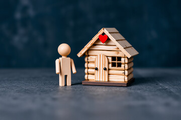 A wooden figure stands beside a miniature wooden house with a red heart. Symbolizes home and love. Simple, crafted design.
