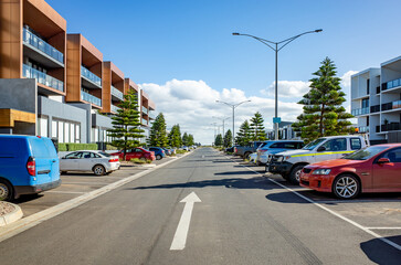 A contemporary urban street featuring modern multi-story apartment buildings, lined with parked...