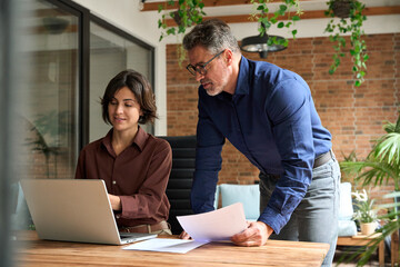 Two busy executive managers working on laptop discussing financial documents at meeting. Middle...