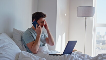 Upset manager speaking mobile phone sitting bed with laptop at hotel closeup