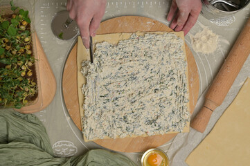 Woman's hand with fork putting cottage cheese with parsley and dill greens on dough. Rolling pin, cutter, egg, green towel and microgreens sprouts on kitchen table. Top view. Home cooking baking puffs