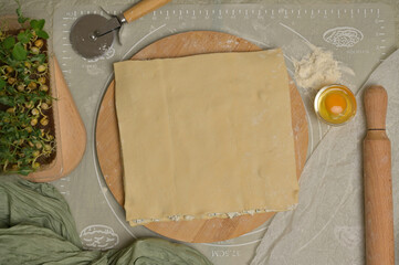 Top view square sheet of dough on wooden board. Rolling pin, cutter, egg, green towel, microgreens sprouts on kitchen table. Home cooking baking puffs