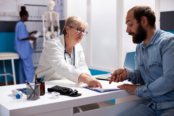 Young male patient completes medical forms with help from senior female doctor, each patiently discussing his information. In clinic, elderly physician shows bearded man where to sign on clipboard.