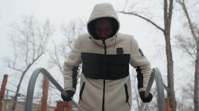 Close-up of young man performing push-up exercise for muscle buildup on outdoor parallel bars during winter, he is wearing a hooded jacket and gloves, with visible breath in the cold air