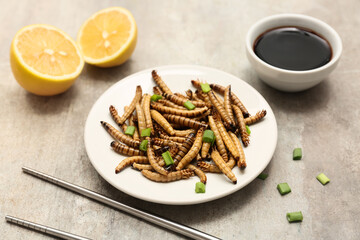 Plate of fried maggots with green onion, lemon and bowl of soy sauce on grey background