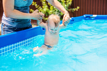 Laughing baby boy playing in a backyard pool, supported by adult hands on a sunny summer day