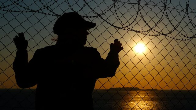 The refugee problem at the border. Silhouette of a desperate refugee at the border with barbed wire against the backdrop of a sea sunset