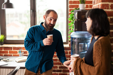 Businessman and woman share conversation near water cooler in brick wall workspace. Male and female employees hold coffee cups combining casual interaction with productive talk in startup office.