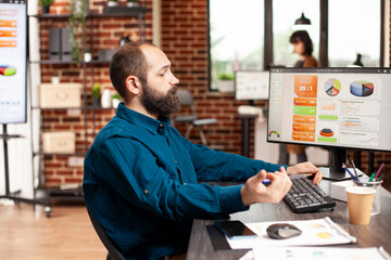 Bearded man in business casual sits at office desk, analyzing financial charts on desktop monitor....