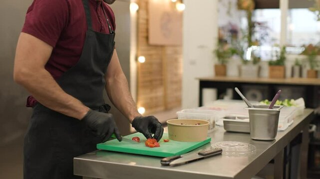 Hispanic chef slices cherry tomatoes and adds them to a compostable bowl with salad, finishing with crushed walnuts. Side view in a cozy takeaway food shop. Natural light, blurred plants. 4k video