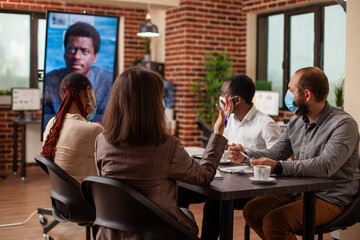 Colleagues attend online training, using digital screen to communicate with remote employer. Team of professionals with face mask in brick wall office, in a virtual meeting, ensuring safety at office.