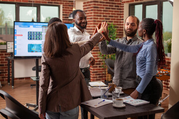 In brick wall office, multiethnic people standing and high fiving over office desk after completion of team discussion. Businesswomen and men celebrating company project deal achievement.