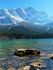 Snow-capped mountains reflect in a serene lake during a clear sunny day near a forest