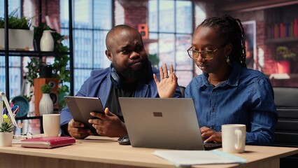 Black woman is working while her partner listens to music next to her, enjoying their time at home together. Freelancer solving her daily tasks on laptop, boyfriend scrolling web. Camera B.