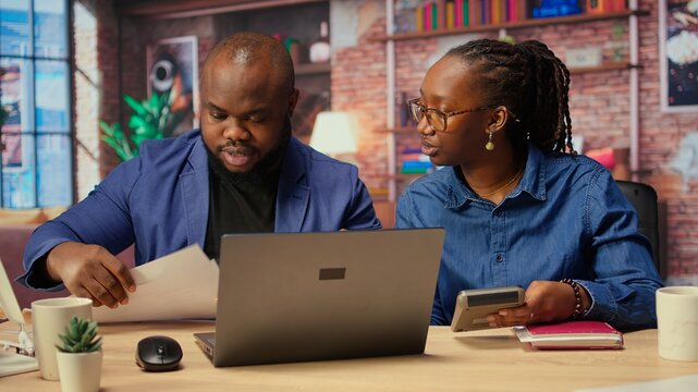 African american man and woman review earnings and revenue reports in their apartment, using calculator and laptop to ensure accurate accounting and financial planning for the tax season. Camera A.