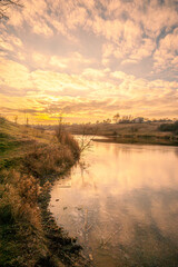 Partially frozen blue lake surrounded by grassy hills with orange grass and yellow reeds against a blue sky with white clouds during sunset.