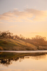 Partially frozen blue lake surrounded by grassy hills with orange grass and yellow reeds against a blue sky with white clouds during sunset.