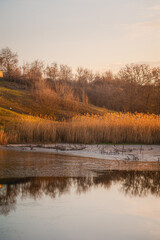 Partially frozen blue lake surrounded by grassy hills with orange grass and yellow reeds against a blue sky with white clouds during sunset.