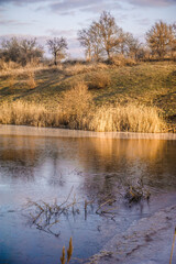 Partially frozen blue lake surrounded by grassy hills with orange grass and yellow reeds against a blue sky with white clouds during sunset.
