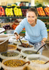 Middle-aged European female picking up olives with scoop from plastic container in grocery store