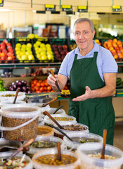 Cheerful old-aged man seller pleasantly offering olives in scoop in large vegetable shop