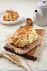 A puff pastry and a croissant are beautifully arranged on a wooden board and a white plate, accompanied by a white teapot, cup, and saucer, creating a cozy breakfast setting.