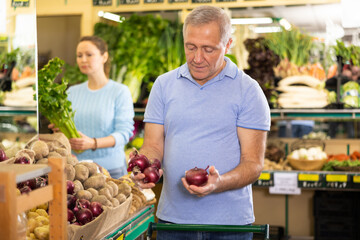 Watchful aged man purchaser taking red onions at the counter in grocery store