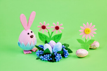 nice toy pink bunny celebrates the holiday of Easter with colorful eggs and flowers on a green background