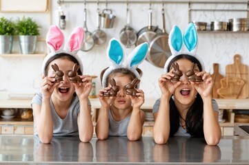 Three cheerful children wearing colorful bunny ears playfully hold up chocolate bunnies in a vibrant kitchen. The atmosphere exudes excitement and creativity during the Easter festivities.