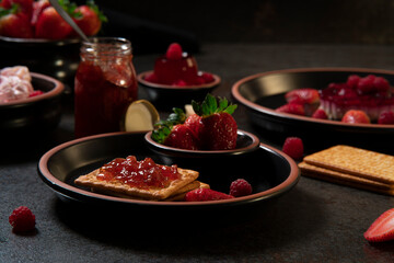 plate with soda crackers with strawberry jam in the foreground against a background of a jam jar and plates with strawberry desserts
