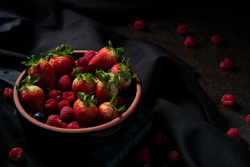 Black ceramic pot filled with red berries, strawberries, raspberries, and blueberries on a black table with a black tablecloth and raspberries