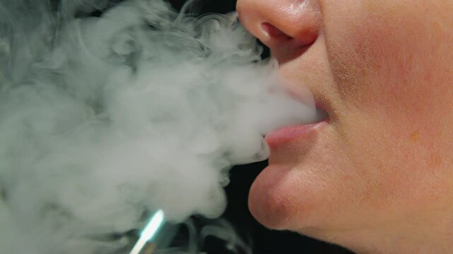 Close-up of mouth of Caucasian woman inhaling thick smoke from vape, on black background. Smoking electronic cigarette. Side view
