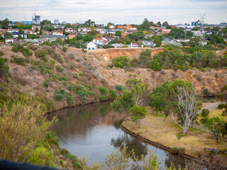 The Maribyrnong River winds through a suburban landscape in Melbourne, Australia, bordered by native trees, grasslands, residential houses, and sloping terrain along the riverbank.