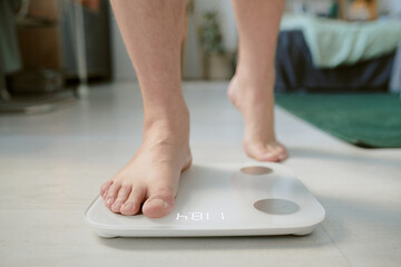 Fototapeta premium Person standing on digital scale with visible weight readout in home setting. Background features household objects and furniture, emphasizing cozy atmosphere