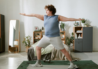 Man performing indoor exercise with outstretched arms, standing on a yoga mat in a well-decorated living room near dumbbells and plants