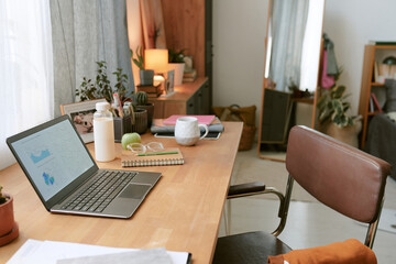 Warm and inviting home office workspace featuring plants, wooden furniture, and organized desk setup. Laptop open with a notebook, water bottle, and lamp complementing ambiance