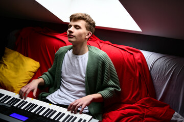 Young man plays synthesizer, musician guy composes track on keyboard sitting on sofa at home