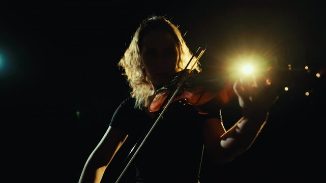 Express yourself by music, woman violin player performing on stage in darkness. Inspired beautiful female silhouette against bright soffit on stage of music-hall or theater, rehearsing before concert