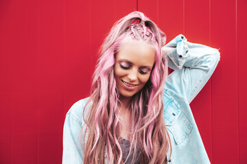 Young beautiful smiling hipster woman in trendy summer jeans shorts with pink hairstyle and makeup. Sexy carefree model posing in the street near red wall. Positive model outdoors