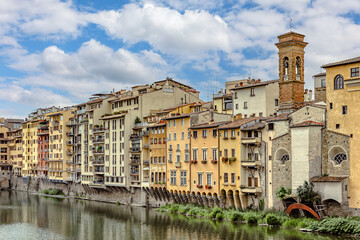 Colorful buildings lining the Arno River in medieval Florence under a blue sky with fluffy clouds