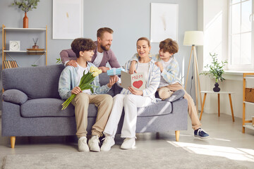 Happy father and clildren, two sons, congratulating mother with International Women Day, birthday or Mother Day. Smiling excited mother getting card, gift box and spring flowers from her loving family