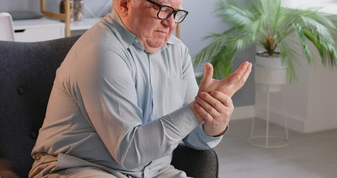 Unhappy and upset elderly man experiencing wrist pain due to arthritis or osteoporosis massages hand on armchair at home. Healthcare, self-care, and coping with rheumatism symptoms.