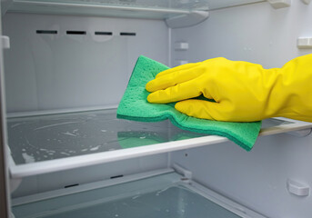 Person cleaning the refrigerator with a cloth. Cleaner wiping the shelves of a clean refrigerator. Hand washer with yellow rubber protective gloves and green sponge.