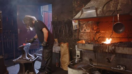 A blacksmith apprentice hammers and is advised by his teacher in the smithy