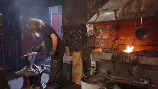 A blacksmith apprentice hammers and is advised by his teacher in the smithy