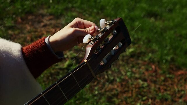 A person adjusts the tuning pegs of a guitar while enjoying a sunny day in a peaceful park. The soft grass and gentle breeze create a serene environment for music.