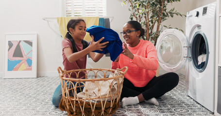 A smiling woman and her daughter take clothes out of the laundry basket and place them into the...