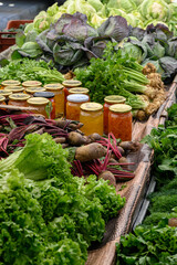 Homemade preserves in glass jars alongside fresh herbs and vegetables at a farmers market. Pickles, jams, and sauces are neatly arranged, creating a vibrant display of natural and organic food.
