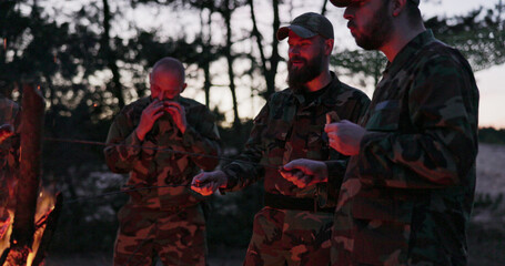 Soldiers in camouflage stand around a fire, engaged in conversation, reflecting on the day's...