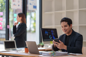 Young happy entrepreneur using a smartphone for online banking, checking emails, browsing social media, ordering food, and making video calls in a bright office
